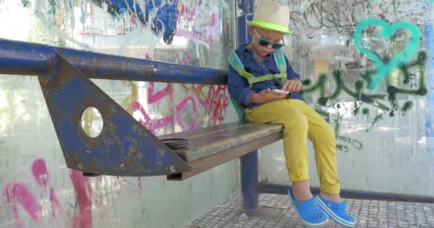 Criança usando telefone inteligente na parada de ônibus grungy — Vídeo de Stock