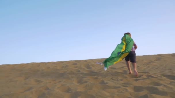 Niño con bandera brasileña corriendo sobre la arena — Vídeo de stock