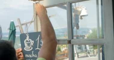 Young man hangs on the door of cafe black board in city of Perea, Greece