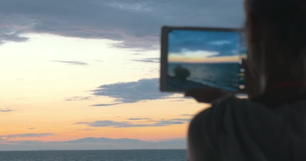 Woman takes photos of the beautiful scenery of the sea and evening sky with her tablet at sunset — Stockvideo