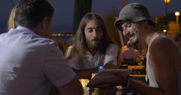 Группа друзей сидит вечером за столом и смотрит видео на планшете, один из мужчин, разговаривает и жестикулирует — стоковое видео