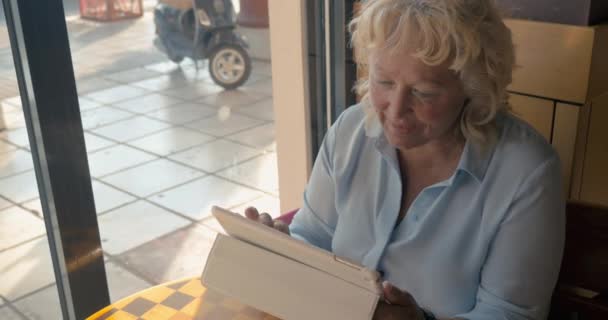 Rijpe vrouw zittend in een café en geniet van haar tablet., buiten het raam de zomer, zonnige dag — Stockvideo