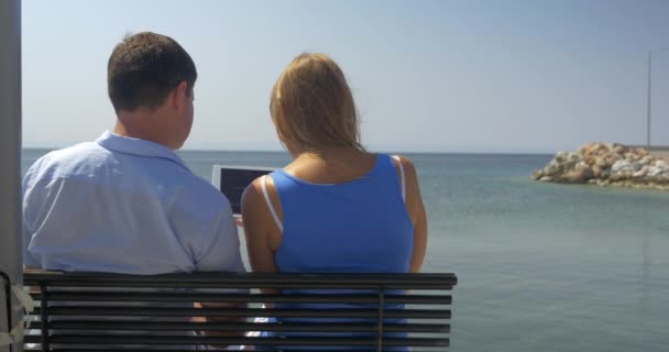 Mujer joven y el hombre están sentados en el banco en la playa en el fondo del horizonte del mar viendo algo en la computadora de la tableta y hablando Pireo, Grecia — Vídeo de stock