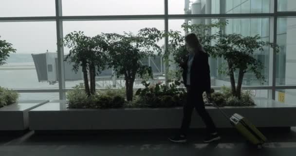 Chica con celda y maleta caminando por la ventana de la terminal del aeropuerto — Vídeos de Stock