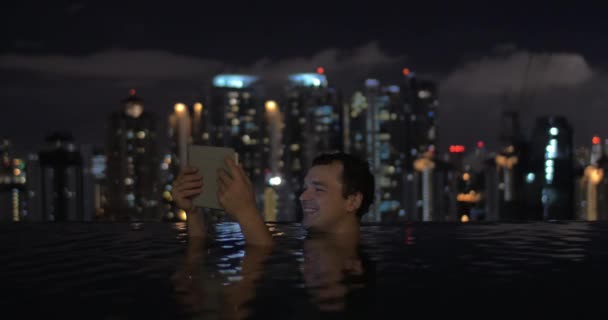 Na piscina no telhado de um hotel em Kuala Lumpur, o homem da Malásia assiste vídeo em tablet e sorri — Vídeo de Stock