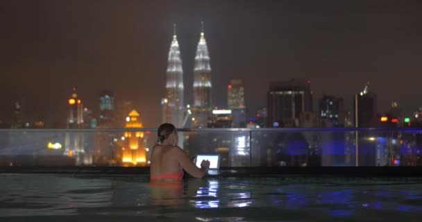 Kadın yüzme havuzunda görünümünü gökdelen çatıda tablet gece şehir manzarası karşı kullanma. Kuala Lumpur, Malezya — Stok video