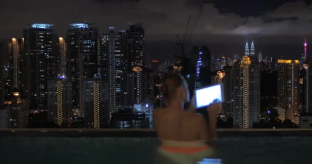Nézd nő beírnak tabletta a medencében a felhőkarcoló tető és az éjszakai város táj. Kuala Lumpur, Malajzia — Stock videók