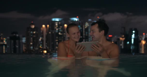 Vista noturna do jovem casal bonito na piscina assistindo em tablet no telhado do arranha-céu e paisagem da cidade. Kuala Lumpur, Malásia — Vídeo de Stock