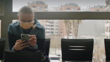 Maskeli kadın bekleme odasında akıllı telefon kullanıyor.