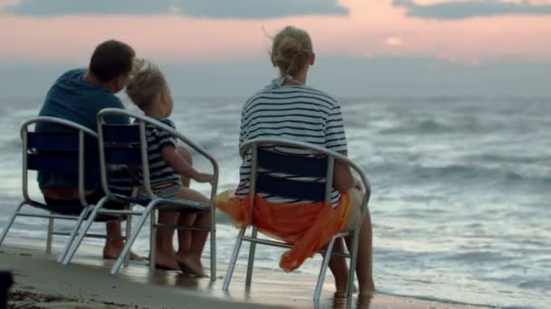 Familia de tres personas sentadas en sillas por mar al atardecer — Vídeos de Stock