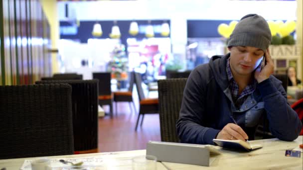 Joven en la cafetería tomando notas mientras habla por teléfono — Vídeo de stock