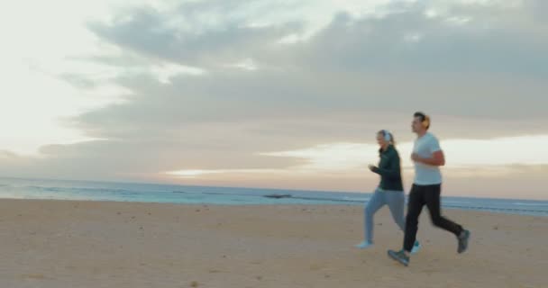 Young couple in headphones running on the beach — Stockvideo