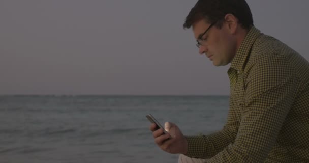 Jeune homme utilisant un téléphone intelligent par la mer dans la soirée — Video