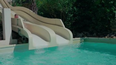 8 yaşında, güneş gözlüğü takan bir kız su kaydırağından aşağı süzülerek havuza giriyor.