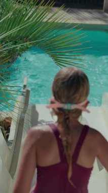 8 yaşında mayo giymiş bir kız su kaydırağından aşağı süzülerek yüzme havuzuna giriyor.