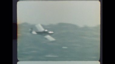 Arka planda şehir manzarası olan uzak bir sahil şeridinde, okyanusun üzerinde alçaktan uçan 8 mm 'lik bir filmde çekilmiş küçük bir uçak.