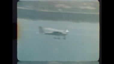 Hafif ışık ve soluk film dokusuyla sakin bir kıyı bölgesinde, uçuşun ortasında yakalanan küçük bir uçak.