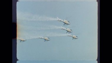 Bir grup helikopter, açık mavi gökyüzünde senkronize akrobatik hareketler yapıyor. 8mm 'lik geçmişe dönük film görüntüsü ve yumuşak bir odak noktası var.