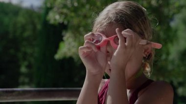 Kırmızı mayo giymiş, güneşli bir günde, arka planda yeşil ağaçlarla pembe yüzme gözlüğü takan genç bir kız.