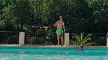 Genç bir çocuk, yeşil ağaçlar ve palmiyelerle çevrili ağır çekimde turkuaz bir havuza atlıyor.