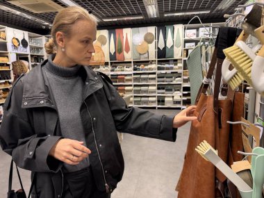 Siyah ceketli bir kadın, rafları eşya dolu bir mağazada deri önlüklerini karıştırıyor.