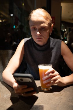 Elinde bir bardak bira tutan ve loş bir kafede tek başına oturan akıllı telefon kullanan bir kadın.