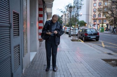 Şehir kaldırımında duran bir kadın telefonuna binaları ve yakınındaki park edilmiş arabalarla bakıyor.