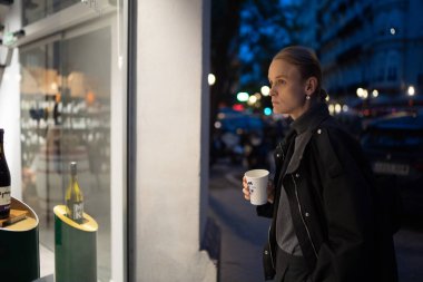 Gece boyunca vitrinlere bakarken elinde kahve fincanı tutan koyu renk ceketli ve gri kazaklı bir kadın.
