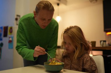 Yemek masasında arkasında duran bir kadın kızına makarna yediriyor.