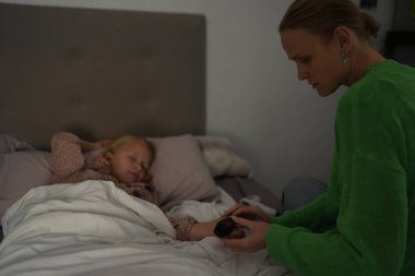 Hasta çocuğu gece yatakta parmak ucu nabız oksimetresi kullanarak kontrol eden bir kadın.