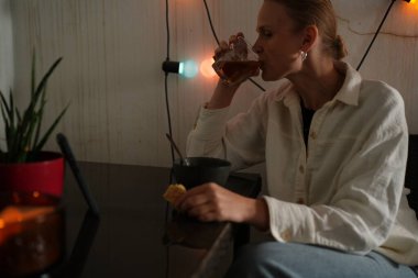 Sıradan giyinmiş bir kadın çay içiyor ve akşam süslenmiş bir masada otururken bir şeyler atıştırıyor.