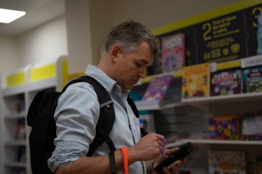 Postanedeki adam elinde turuncu tasmayla kitap raflarının yanında dijital tablet kullanıyor.