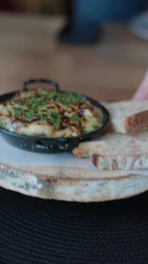 Eritilmiş fırınlanmış peynirli sıcak tava, ahşap tahtada kızartılmış ekmek dilimleriyle servis edilen otlarla kaplı.