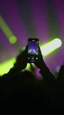 Silüet eller akıllı telefonu tutuyor canlı yeşil ve mor sahne ışıklandırmasıyla canlı konser kaydediyor.