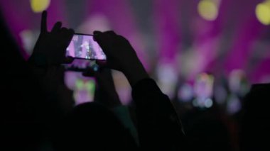 Renkli sahne ışıkları ve bulanık kalabalıkla akıllı telefon kayıt konseri tutan ellerin yakın görüntüsü