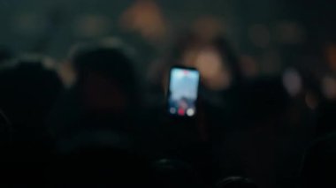 Karanlık kalabalık ve sahne parıltısıyla çevrili akıllı telefon kayıt konserinin yakın görüntüsü yoğun müzik enerjisini yakalıyor