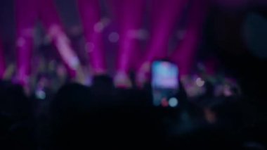 Konser kalabalığının dışında, mor sahne ışıkları altında yükseltilmiş akıllı telefonlar yüksek enerjili canlı müzik atmosferi yaratıyor.