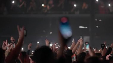 Taraftarlar akıllı telefonları ve elleri kaldırarak sahnede güçlü bir konser anını yakalıyorlar. Flaş flaşlı ve dinamik atmosferli.