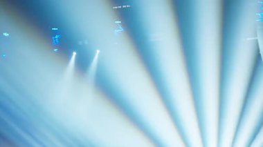 Yoğun mavi sahne ışıkları karanlık mekan boyunca geniş kirişler yansıtıyor. Canlı performansın ve konser deneyiminin ölçeğini vurguluyor.