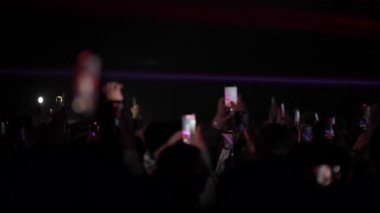 Karanlık konser salonu atmosferinde yükseltilmiş akıllı telefonlar ve parlak sahne ışıklarıyla kalabalık bir film gösterisi.
