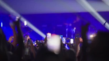 Konser izleyicileri, mavi ışıklandırmalı enerjik canlı performans sırasında el kaldırıyor, akıllı telefonlar çalıyor.