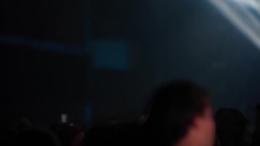 Karanlık kalabalıktaki insanlar sahneyi yavaş çekimde izliyorlar parlak spot ışıkları sisi keserken yoğun bir canlı müzik ortamı yaratıyor.