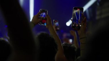 Arkadan, bir hayran kitlesi canlı müzik konseri kaydetmek için akıllı telefonları tutuyor. Sahne, telefon ekranlarına yansıyan parlak mavi ışıklarla aydınlatılıyor. Modern, enerjik bir olay ortamı.