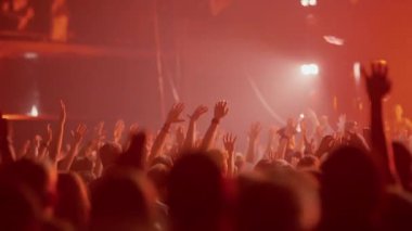 Elleri havada dans eden ve bir müzik festivalinde tezahürat yapan büyük, enerjik bir kalabalık. Sahne canlı kırmızı ışık ve duman içinde, dinamik ve canlı bir atmosfer yaratıyor..