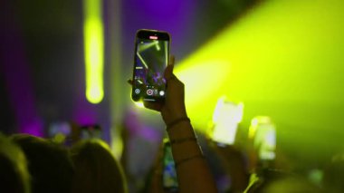 Kalabalıktaki bir hayran canlı konser kaydetmek için bir akıllı telefonu soyuyor. Telefon ekranı, performansı parlak sarı ve mor sahne ışıkları altında gösteriyor. Diğer telefonlar görünür..