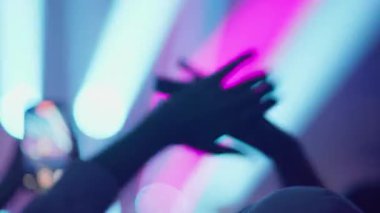 Kalabalığın içinde akıllı telefondan konser kaydeden bir kişinin silueti. Pembe ve mavi sahne ışıklarının canlı, bulanık arka planına karşı ellerini kaldırır, el sallar ve müziği alkışlar..