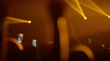 Bir müzik festivalindeki kalabalığın siluetinin videosu. Dinamik altın ve kırmızı sahne ışıkları enerjik bir atmosfer yaratırken hayranlar şovu kaydetmek için telefonları tutuyor. Etkileyici bir bakış açısı..