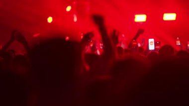 Canlı müzik konseri veya festivalinde dans eden ve tezahürat yapan siluetli insanların büyük bir kalabalığının görüntüsü. Sahne yoğun kırmızı ışıkla yıkanırken eller ve telefonlar havada..