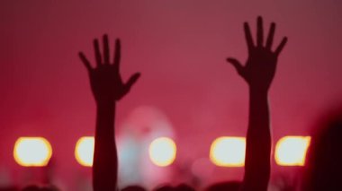 Canlı müzik konserinde ya da festivalde elleri havada bir kalabalığın silueti. Kırmızı sahne ışıkları ve duman atmosferik ve enerjik bir kutlama ve ibadet sahnesi oluşturur..