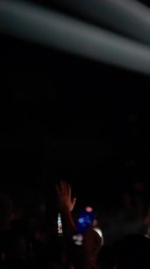 Canlı müzik konserinde kalabalığın arkasından dikey çekim. Sahne ışıkları dumanlı, karanlık mekanı süpürürken hayranlar kaydetmek için telefonları engelliyor. Heyecanla el kaldırılır. Enerjik atmosfer.